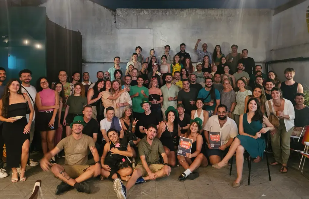 A big, smiling crowd at a Drink’n’Talk night, some in green hats, two people holding up Drink’n’Talk signs.
