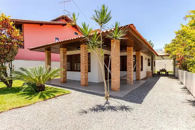 A single-storey house with brick pillars, a porch with a hammock, a palm and a sunny gravel yard.